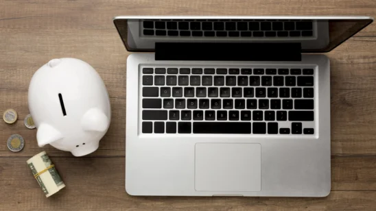 Notebook aberto ao lado de um cofrinho de porquinho branco, moedas e um maço de dinheiro sobre uma mesa de madeira, representando o tema "salário especialista em marketing digital".