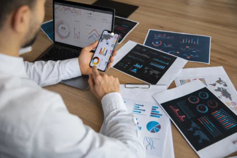 Profissional de análise de dados revisando gráficos e dashboards em um laptop e smartphone, com diversas representações visuais de dados espalhadas pela mesa, exemplificando as habilidades necessárias para a análise de dados.