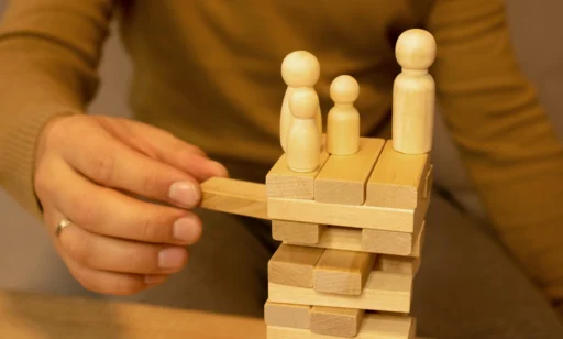 Mão de uma pessoa mexendo em blocos de madeira empilhados, com figuras de pessoas esculpidas no topo dos blocos. A imagem representa o conceito de equilíbrio e desafios na gestão de equipes e problemas no rh.