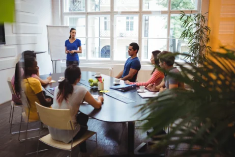 Grupo de profissionais reunidos em uma sala de conferência com uma profissional de RH conduzindo uma reunião, exemplificando as funções no RH no dia a dia.