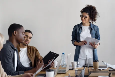 Grupo de talentos internos em uma reunião de trabalho. Uma mulher, em pé, segura um tablet enquanto escuta dois homens sentados à mesa. Eles discutem documentos e trabalham em laptops. A mesa está organizada com copos, cadernos e garrafas de água.